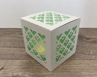 May include: A white, cube-shaped paper lantern with geometric cutouts. The cutouts reveal a green interior, creating a contrast with the white exterior. The lantern sits on a wooden surface.