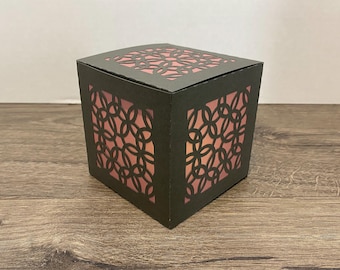 May include: A dark gray paper lantern with a geometric cutout design. The lantern has a square shape with a pink interior, creating a warm glow. The design is a repeating pattern of interlocking circles.