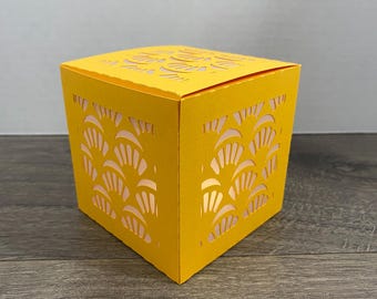 May include: A yellow paper box with a geometric fan-like pattern cut into the sides. The box is cube-shaped and sits on a wooden surface. The interior of the box is a lighter color, creating a contrast with the yellow exterior.