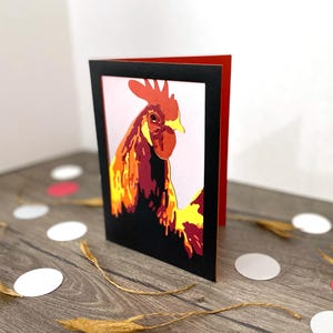 May include: A greeting card featuring a stylized rooster illustration. The artwork uses shades of red, orange, and yellow against a black frame, with a red interior. The card is standing on a wooden surface with decorative elements.