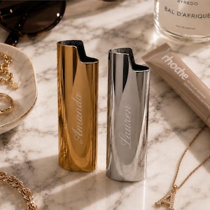 May include: Two metallic lighter cases standing upright. The left case is gold-toned with the name Amanda engraved in script. The right case is silver-toned with the name Lauren engraved in script. Both cases feature a smooth, reflective finish.