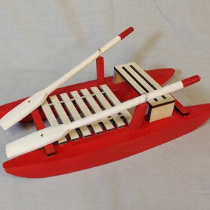 Può includere: Una barca giocattolo di legno rossa con due remi bianchi. La barca ha un ponte a doghe bianche e un piccolo sedile di legno.