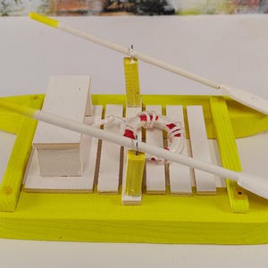 Könnte beinhalten: Ein gelbes Holzspielzeugfloß mit zwei weißen Rudern und einem roten und weißen Rettungsring. Das Floß hat eine weiße Holzplattform mit einer kleinen weißen Holzkiste.