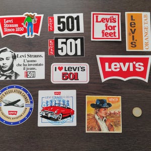 Puede incluir: Una colección de pegatinas y parches vintage de la marca Levi's. Las pegatinas presentan varios diseños, incluido el logotipo de Levi's, el número 501 y la frase "Levi's for feet".