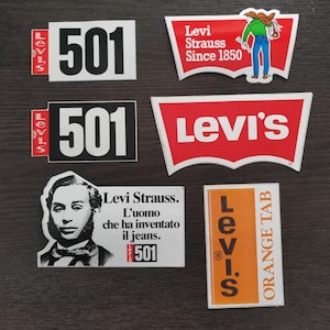 Może przedstawiać: Kolekcja sześciu prostokątnych i trapezoidalnych naklejek z brandingiem Levi's. Wzory obejmują liczbę 501, logo Levi Strauss z postacią, logo Levi's batwing, portret Levi Straussa z włoskim tekstem oraz etykietę typu orange tab.