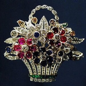 May include: A detailed brooch in the shape of a basket overflowing with colorful gemstones. The basket is silver-toned, with a green band at the base. The gemstones are various colors, including red, blue, and clear, arranged to resemble flowers.