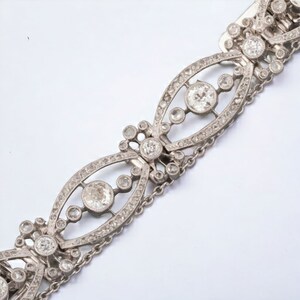 May include: A silver bracelet with an intricate design. The bracelet features multiple oval-shaped links, each adorned with small, sparkling stones and larger, round gemstones. Delicate chain links connect the decorative sections.