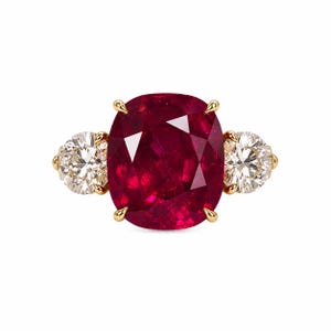Puede incluir: Un anillo de oro con un gran rubí rojo intenso de talla cojín en el centro, flanqueado por dos diamantes blancos redondos de talla brillante. Las piedras están engastadas en garras de oro amarillo, creando un diseño clásico de tres piedras.