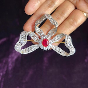 May include: A silver-toned brooch in a bow shape, adorned with sparkling clear crystals and a central oval-cut red gemstone. The brooch has a pin closure on the back and is held against a dark purple background.