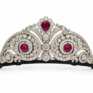 May include: A silver-toned tiara featuring a central pear-shaped ruby surrounded by diamonds, flanked by two circular ruby cabochons set within swirling diamond-encrusted scrolls. The piece is mounted on a thin black band with intricate metalwork.