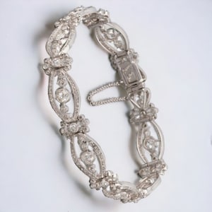 May include: A silver bracelet with an ornate design. The bracelet features oval links with a central diamond and smaller surrounding stones. The clasp is visible, along with a safety chain. The bracelet is likely made of sterling silver or a similar metal.