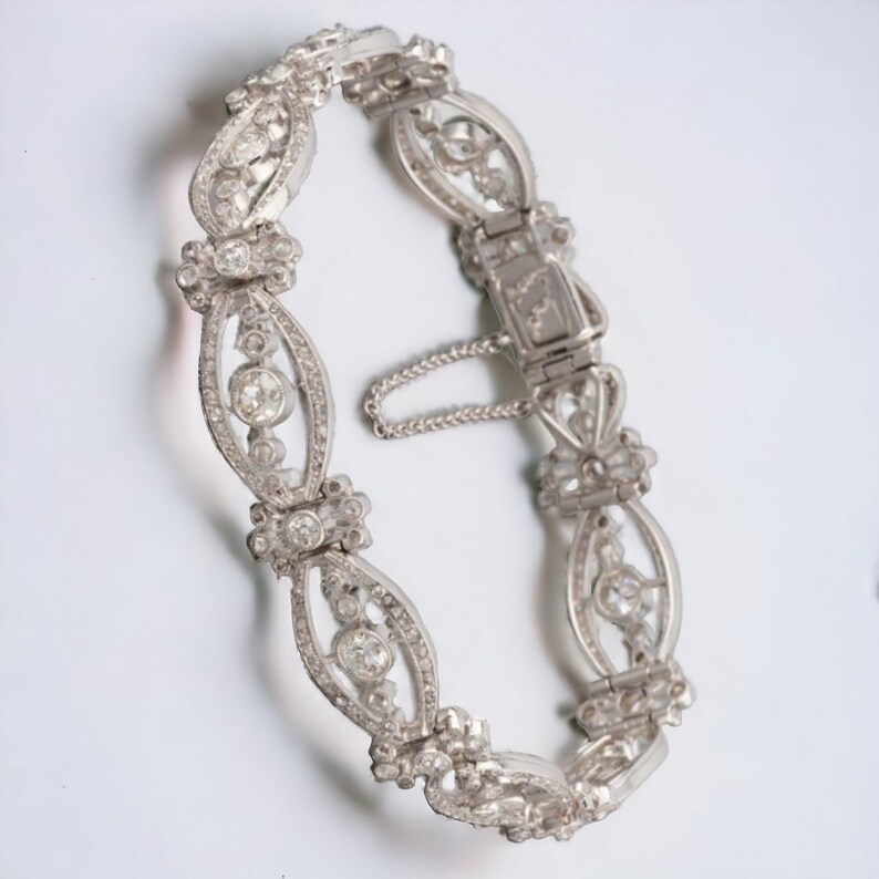 May include: A silver bracelet with an ornate design. The bracelet features oval links with a central diamond and smaller surrounding stones. The clasp is visible, along with a safety chain. The bracelet is likely made of sterling silver or a similar metal.