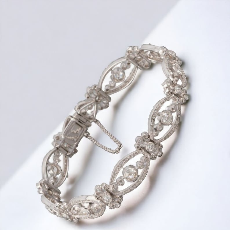 May include: An ornate silver bracelet with a delicate, openwork design. The bracelet features a series of oval links, each adorned with sparkling gemstones. The clasp is visible, and the overall aesthetic is elegant and timeless.