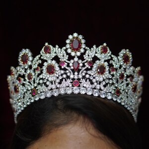 May include: A silver-toned crown with numerous clear and red gemstones. The crown features a repeating pattern of oval red stones surrounded by smaller clear stones. The crown is set against a dark background.
