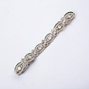 May include: A silver-toned bracelet with an ornate design. The bracelet features a series of oval-shaped links, each encasing a central, round, clear stone, surrounded by smaller, sparkling stones. The links are connected by delicate, silver-toned metalwork.