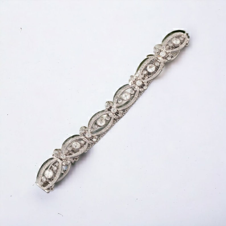 May include: A silver-toned bracelet with an ornate design. The bracelet features a series of oval-shaped links, each encasing a central, round, clear stone, surrounded by smaller, sparkling stones. The links are connected by delicate, silver-toned metalwork.
