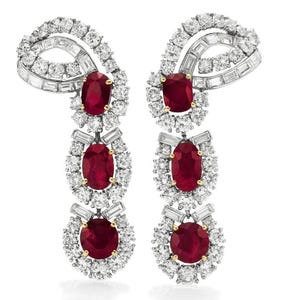 May include: A pair of dangle earrings with a silver setting and three red ruby stones in each earring. The ruby stones are surrounded by smaller diamonds.