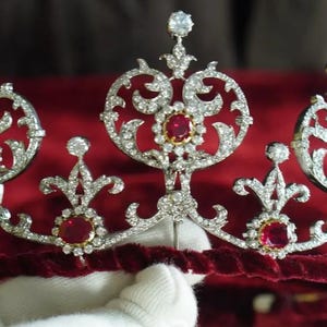 Może przedstawiać: Ozdobna srebrna tiara z błyszczącymi, przezroczystymi kamieniami szlachetnymi i żywymi czerwonymi klejnotami. Tiara ma aksamitną opaskę i jest osadzona na bogatym czerwonym tle. Projekt zawiera wzory kwiatowe i okrągłe.