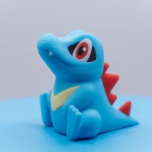 May include: A blue and red figurine of a Pokemon character with a yellow stripe on its chest.