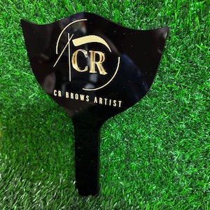 Pode incluir: Um estêncil de sobrancelha de acrílico preto com uma alça, com letras douradas que dizem "CR BROWS ARTIST" e um logotipo "CR" estilizado. O estêncil é exibido em uma cama de grama artificial verde.