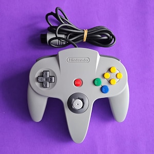 Puede incluir: Un mando gris de Nintendo 64 con cable negro y botones rojos, verdes, azules y amarillos. El mando tiene una cruceta, un joystick y el logotipo de Nintendo.