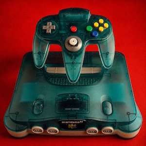 Peut inclure: Une console de jeu vidéo Nintendo 64 bleu sarcelle translucide avec sa manette assortie. La console présente un logo NINTENDO 64 en façade, quatre ports de manette et un emplacement pour extension mémoire. La manette inclut une croix directionnelle grise, un bouton start rouge, des boutons C jaunes, ainsi qu'un bouton A vert et un bouton B bleu.