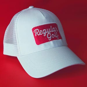 Op de afbeelding: Witte truckerpet met een mesh achterkant en een gebogen klep. De pet heeft een rood rechthoekig embleem met de tekst "Regular Golf" in witte letters. De achtergrond is rood.