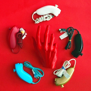 Pode incluir: Uma coleção de controles Wii em várias cores, incluindo branco, vermelho, azul e verde-oliva, dispostos sobre um fundo vermelho vibrante. Uma escultura de mão vermelha está no centro. Os controles estão conectados a cabos.