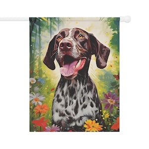 Op de afbeelding: Een decoratieve tuin vlag met een vrolijke Duitse Staande Hond met een bruin-witte vacht, tegen een kleurrijke bloemen achtergrond. De hond heeft een roze tong en is omringd door bloemen.