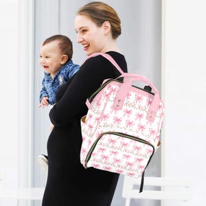 May include: A white and pink diaper bag with a bow and floral pattern. The bag has pink straps and a top handle. The bag is being carried by a person wearing a black dress. A baby is being held by the person.