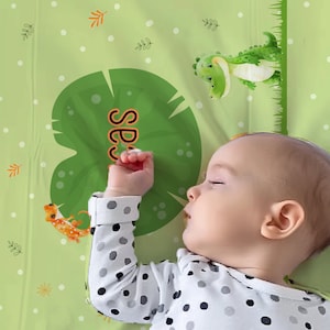 May include: A baby sleeping on a green and white patterned sheet with a green lily pad with the word "Sleepy" on it. There is a green alligator and a lizard on the sheet.