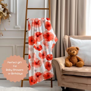 May include: A white throw blanket with a red poppy floral pattern. The blanket is draped over a wooden ladder and a chair with a teddy bear.