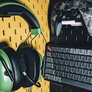 May include: A green and black gaming headset, a black and gray keyboard, and a camouflage-patterned video game controller are mounted on a yellow pegboard wall.