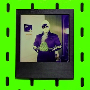 May include: A framed photograph featuring a person in a dark suit and mask holding a pineapple. The image has a surreal, stylized aesthetic with a green and purple color scheme. The frame is black, set against a bright green background.