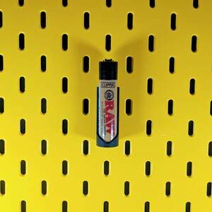May include: A black lighter with a RAW rolling papers design on a yellow pegboard.