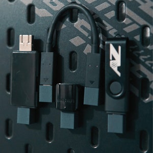 Puede incluir: Primer plano de cuatro dispositivos electrónicos negros con cables. Los dispositivos están montados en una pared gris oscura con un patrón repetitivo. El dispositivo de la derecha tiene el texto "NZ".