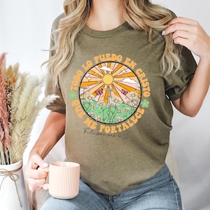 May include: A green t-shirt with a circular graphic featuring a sun, flowers, and butterflies. The graphic includes the text "Todo lo puedo en Cristo que me fortalece" and "Philippians 4:13".
