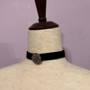 May include: A black velvet choker necklace with a decorative silver button. The button has an ornate, floral design. The choker is displayed on a mannequin with a wooden neck and head. The background is a solid light purple.