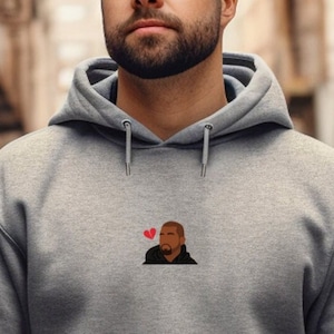 Op de afbeelding: Een grijze hoodie met een voorzak en een capuchon. De hoodie heeft een klein geborduurd ontwerp van een persoon met een rood hart boven hun hoofd. De persoon draagt een zwarte jas. De hoodie is gemaakt van een zacht materiaal.