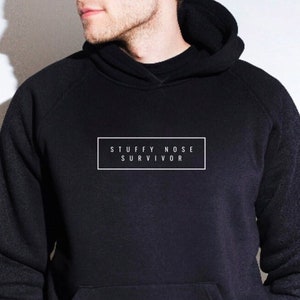 May include: Black hoodie with a front pocket and a hood. The text "STUFFY NOSE SURVIVOR" is printed in a white rectangle on the front. The person is wearing tan cargo pants.