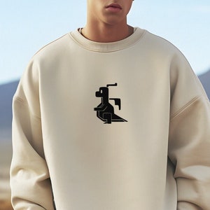 May include: Cream-colored sweatshirt with a black graphic design on the chest. The design appears to be an abstract representation of a creature or object. The sweatshirt has a crew neck and long sleeves. The model is wearing khaki pants.