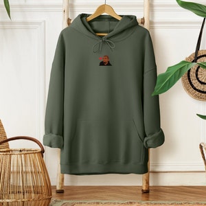 Op de afbeelding: Olijfgroene hoodie met een voorzak en een capuchon met trekkoord. De hoodie heeft een kleine afbeelding van een persoon met een rood hart boven hun hoofd. Hij hangt aan een houten rek.