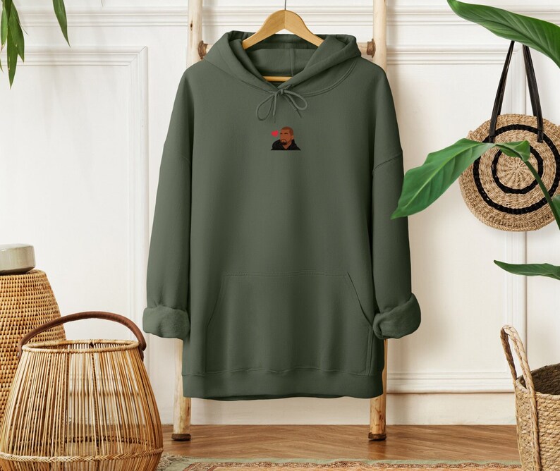 Op de afbeelding: Olijfgroene hoodie met een voorzak en een capuchon met trekkoord. De hoodie heeft een kleine afbeelding van een persoon met een rood hart boven hun hoofd. Hij hangt aan een houten rek.
