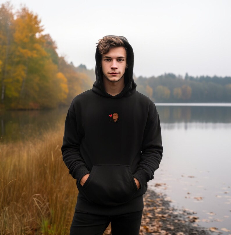 Op de afbeelding: Zwarte hoodie met een voorzak en capuchon. De hoodie heeft een klein geborduurd ontwerp van twee gezichten en een rood hart. De persoon staat voor een meer met herfstbladeren.