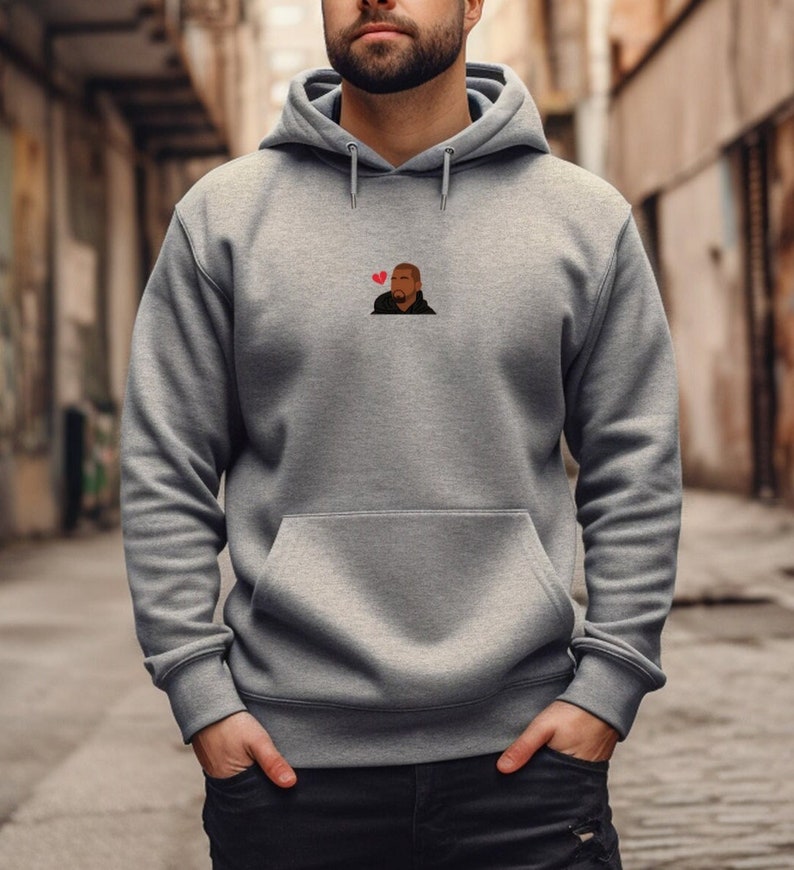 Op de afbeelding: Een grijze hoodie met een voorzak en een capuchon. De hoodie heeft een klein geborduurd ontwerp van een persoon met een rood hart boven hun hoofd. De persoon draagt een zwarte jas. De hoodie is gemaakt van een zacht materiaal.