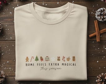 Könnte beinhalten: Ein cremefarbenes T-Shirt mit festlichem Weihnachtsdesign und dem Text "HOME FEELS EXTRA MAGICAL This season." Das Design umfasst Lebkuchenmänner, Weihnachtsbäume und andere Feiertags-Icons. Das Shirt wird auf einer Holzoberfläche präsentiert.