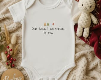 Könnte beinhalten: Weißer Baby-Body mit dem Text "Dear Santa, I can explain... I'm new" und kleinen Illustrationen. Ein gehäkeltes Spielzeugkaninchen und ein Holzschild mit der Aufschrift "Baby" sind ebenfalls im Bild.