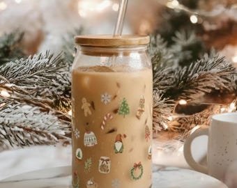 Könnte beinhalten: Ein klares Glas mit einem Bambusdeckel und einem durchsichtigen Strohhalm, gefüllt mit einem hellbraunen Getränk und Eiswürfeln. Das Glas ist mit weihnachtlichen Aufklebern verziert. Im Hintergrund befindet sich eine weiße Tasse.