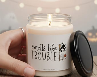 Könnte beinhalten: Eine brennende Kerze in einem durchsichtigen Glasbehälter mit schwarzem Deckel, beschriftet mit "Smells like Trouble" und einer Geistergrafik. Die Kerze steht auf einer Holzoberfläche, mit einer Hand, die das Glas hält. Der Hintergrund zeigt einen Vollmond und Halloween-Dekorationen.