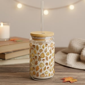 May include: A clear glass tumbler with a bamboo lid and a clear straw. The tumbler is decorated with a pattern of pumpkins, leaves, and mushrooms in shades of orange, yellow, and brown. The tumbler is sitting on a wooden surface.